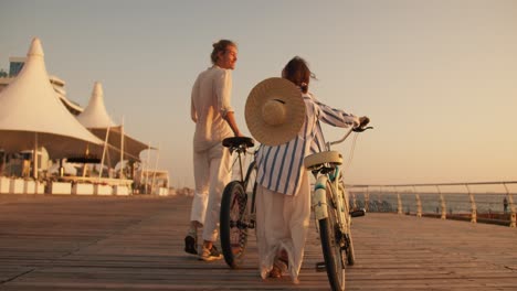 Rückansicht-Eines-Mannes-Und-Eines-Mädchens-In-Leichter-Strandkleidung,-Die-Mit-Ihren-Fahrrädern-Bei-Sonnenaufgang-Im-Sommer-Am-Mit-Brettern-Bedeckten-Strand-Entlang-Des-Meeres-Spazieren