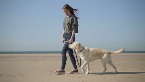 smiling young woman talking to dog while walking on seashore.