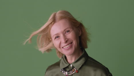 Mujer-Madura-Rubia-Con-Camisa-Verde-Sonriendo-A-La-Cámara-Mientras-El-Viento-Mueve-Su-Cabello-Y-Camisa-Sobre-Fondo-Verde