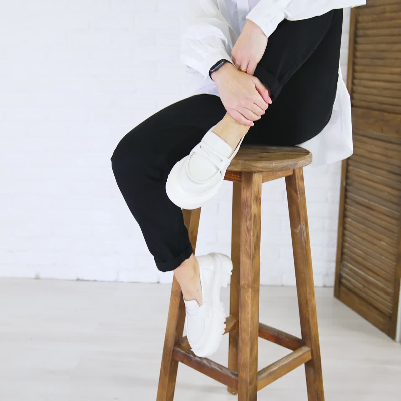 Stylish white loafers on the feet of a lady sitting on high stool. Model touches the shoes then changes her position. White wall backdrop