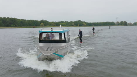 hydro foilers surfing wave behind watercraft driven by blond girl filmed with drone from front close up with trees on riverbank