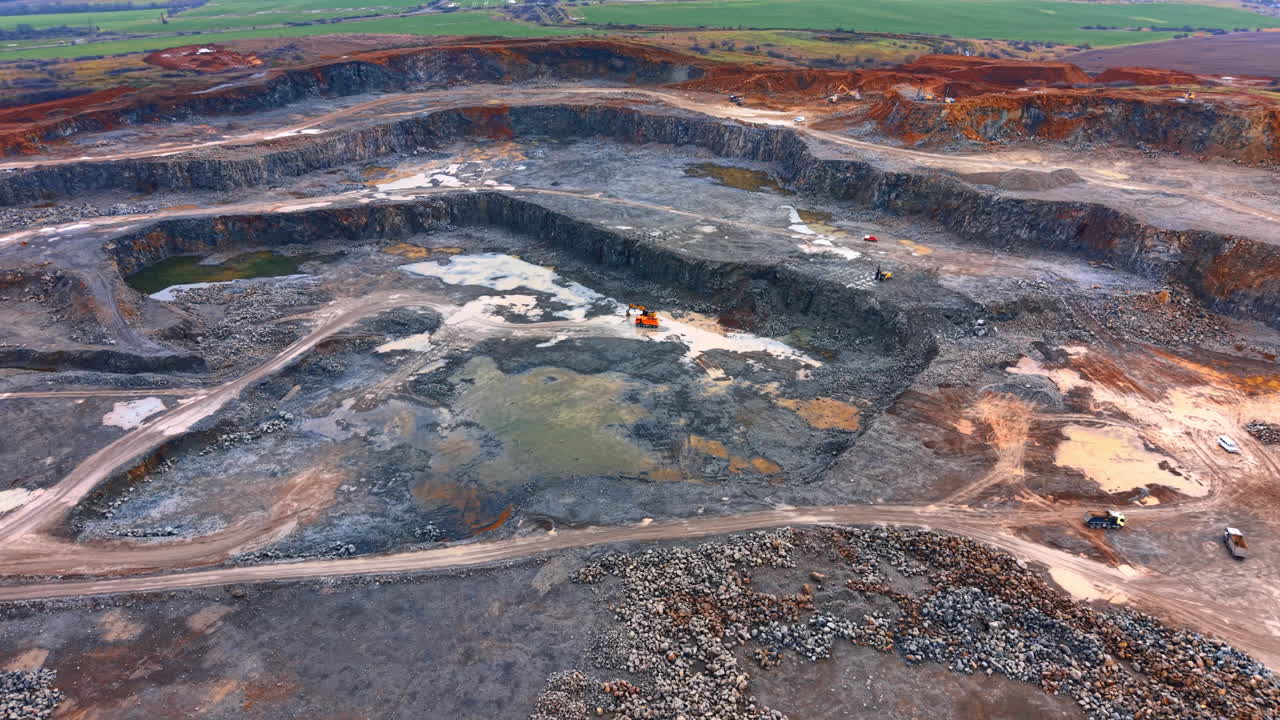 Pit and trucks at excavation site. A large excavation site with trucks working during the day. The ground is uneven with some water puddles