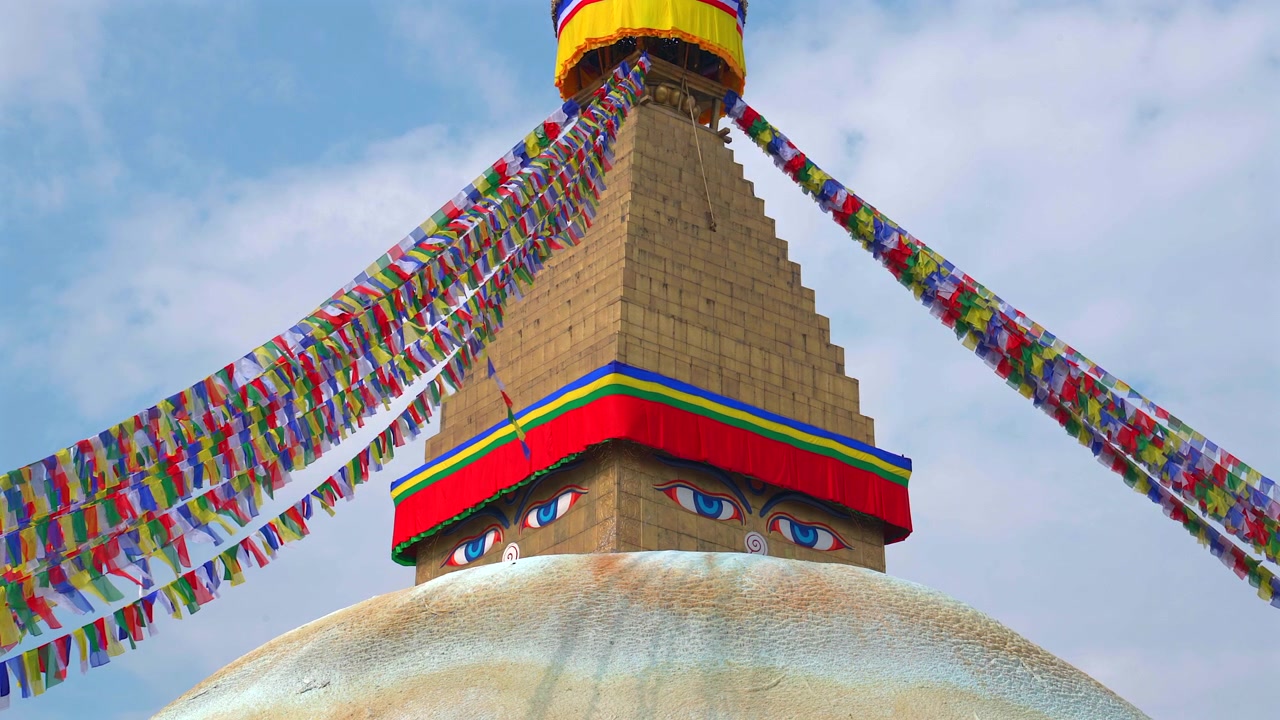 Un'inquadratura statica a media angolazione bassa che si concentra sulla guglia dorata e sugli occhi sacri della saggezza dello Stupa di Boudhanath, circondato da bandiere di preghiera colorate e sventolanti a Kathmandu, Nepal