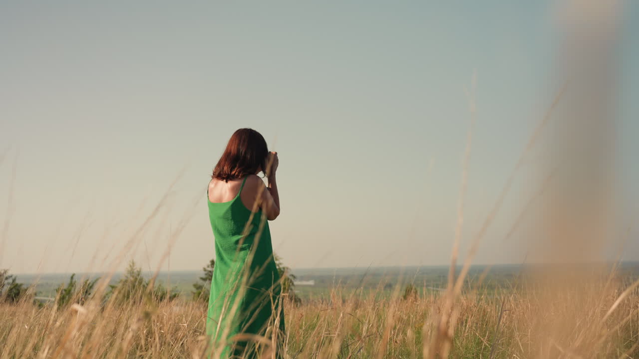woman in green dress steps forward in golden field raising camera to capture scenic view of vast open landscape under calm sky during golden hour creating peaceful outdoor moment