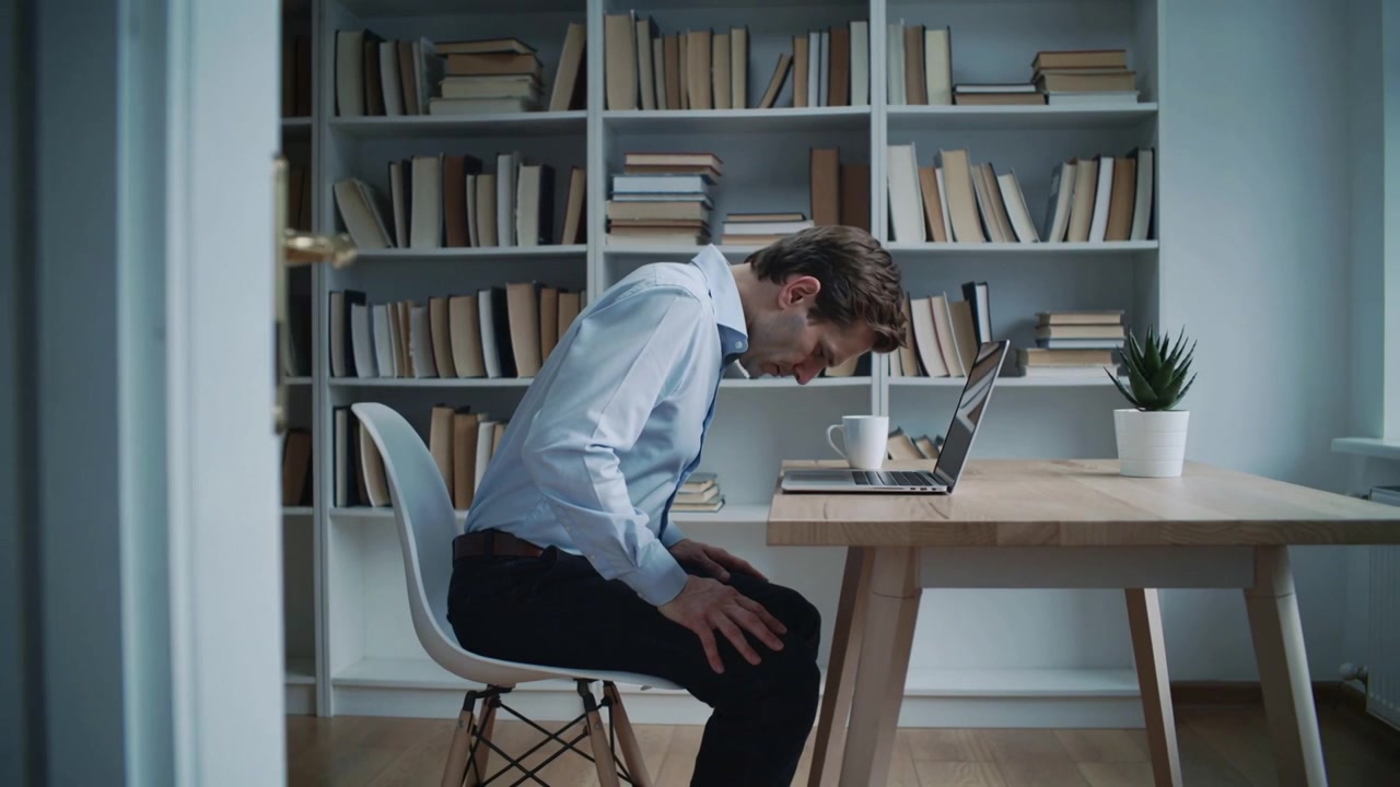 Leaning man in light blue dress shirt tilting forward at home office desk, focusing on laptop