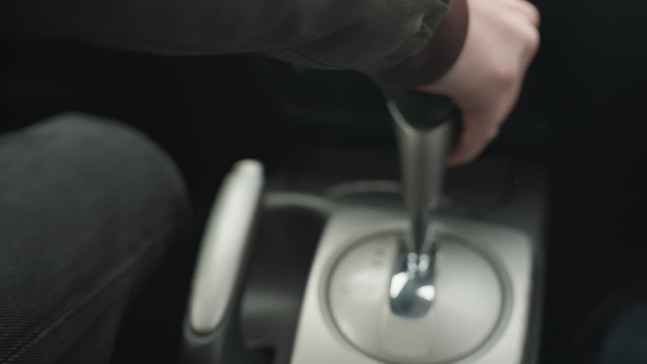 rear view of driver pushing gear lever into park then lifting hand brake lever on center console inside car cabin close up showing safe parking procedure with grip metal finish detail