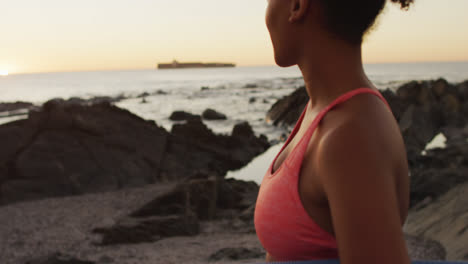 Portrait-of-african-american-woman-holding-yoga-mat-standing-on-the-rocks-near-the-sea-during-sunset