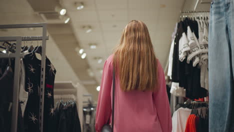 back view of lady carrying black bag walking through a clothing store as she touches a black top and moves forward, the store is well-lit, with neatly arranged clothes on racks on both sides