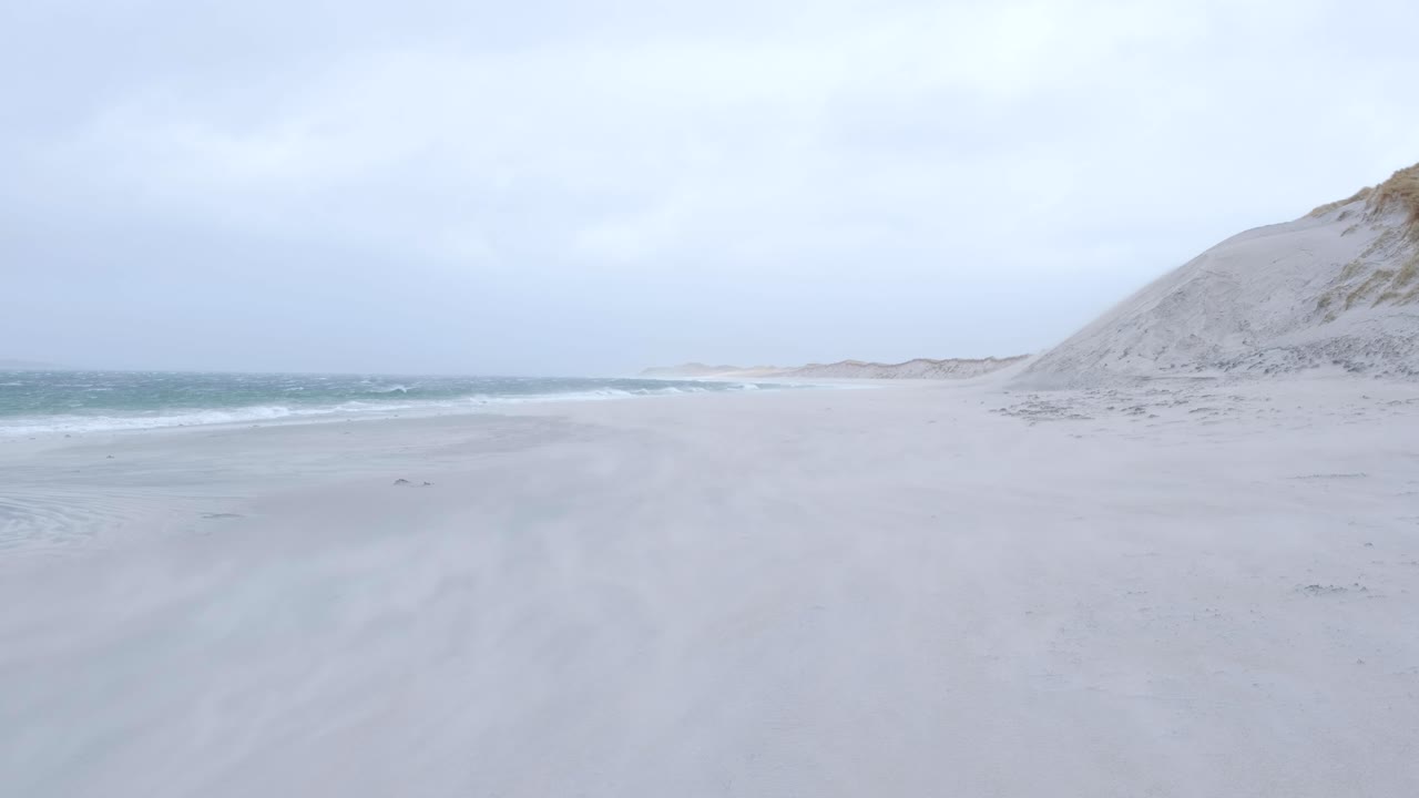 야생 바람이 부는 날씨 동안 모래 폭풍은 스코틀랜드 서부의 외 헤브리드 제도의 베르네레이에 있는  모래 해변의 표면을 가로질러 불고 있습니다.