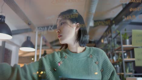 focused asian casual businesswoman brainstorming, making notes on glass wall at night in office