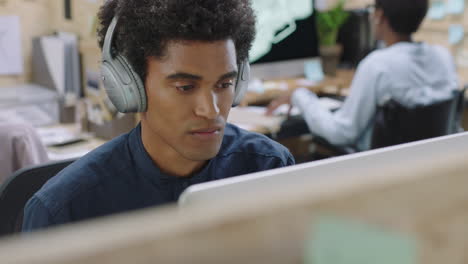 young-mixed-race-businessman-using-computer-working-on-business-project-browsing-online-listening-to-music-enjoying-drinking-juice-wearing-headphones-in-relaxed-office-workplace