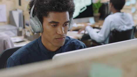 young mixed race businessman using computer working on business project browsing online listening to music enjoying drinking juice wearing headphones in relaxed office workplace