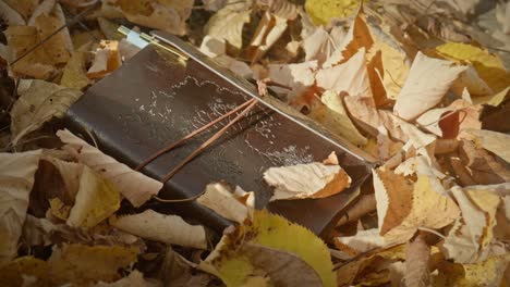 Brown-leather-diary-journal-lays-on-ground,-golden-autumn-leaves-fall