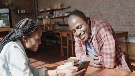 Feliz-Pareja-Afroamericana-Mirando-El-Teléfono-Inteligente-Y-Hablando-En-La-Mesa-En-La-Cafetería,-Cámara-Lenta