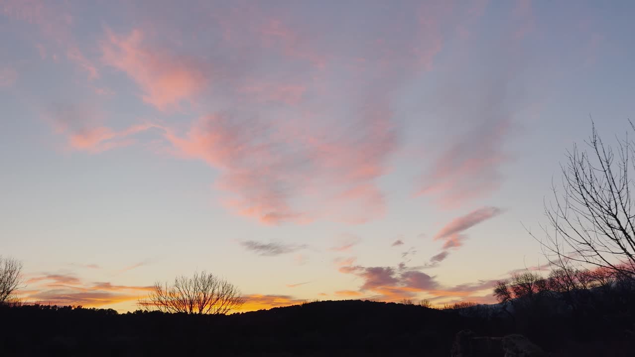 Sunset scene with dark mountain silhouettes and bare tree tops in the foreground. Warm orange light appears in small clouds near the horizon, while the sky shows soft blue and red tones