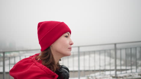 primer plano de una mujer caminando cerca de una barandilla de hierro con gorra roja y chaqueta con auriculares alrededor del cuello, el fondo presenta un puente borroso y un asentamiento urbano en una atmósfera de invierno niebla y cubierta de nieve