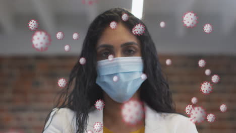 animación de células de covid-19 flotando sobre el retrato de una mujer biracial con máscara facial en la oficina