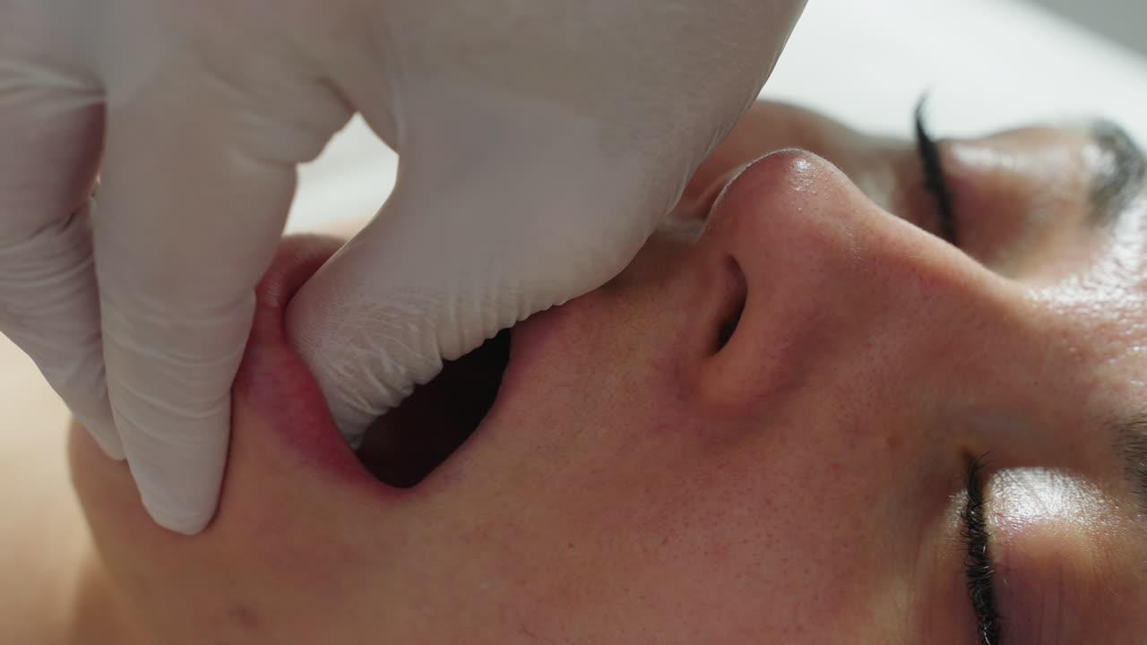 Close up of customer covered with clinical blanket eyes closed and mouth open in relaxed manner as wellness technician wearing gloves dips fingertips into mouth while massaging jawline in spa session