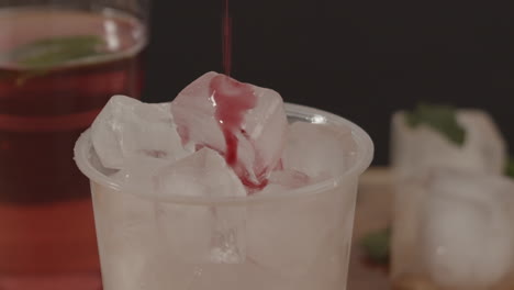 foto de estudio de jarabe rojo vertiendo en un vaso de plástico lleno de hielo con limonada y hielo en segundo plano.