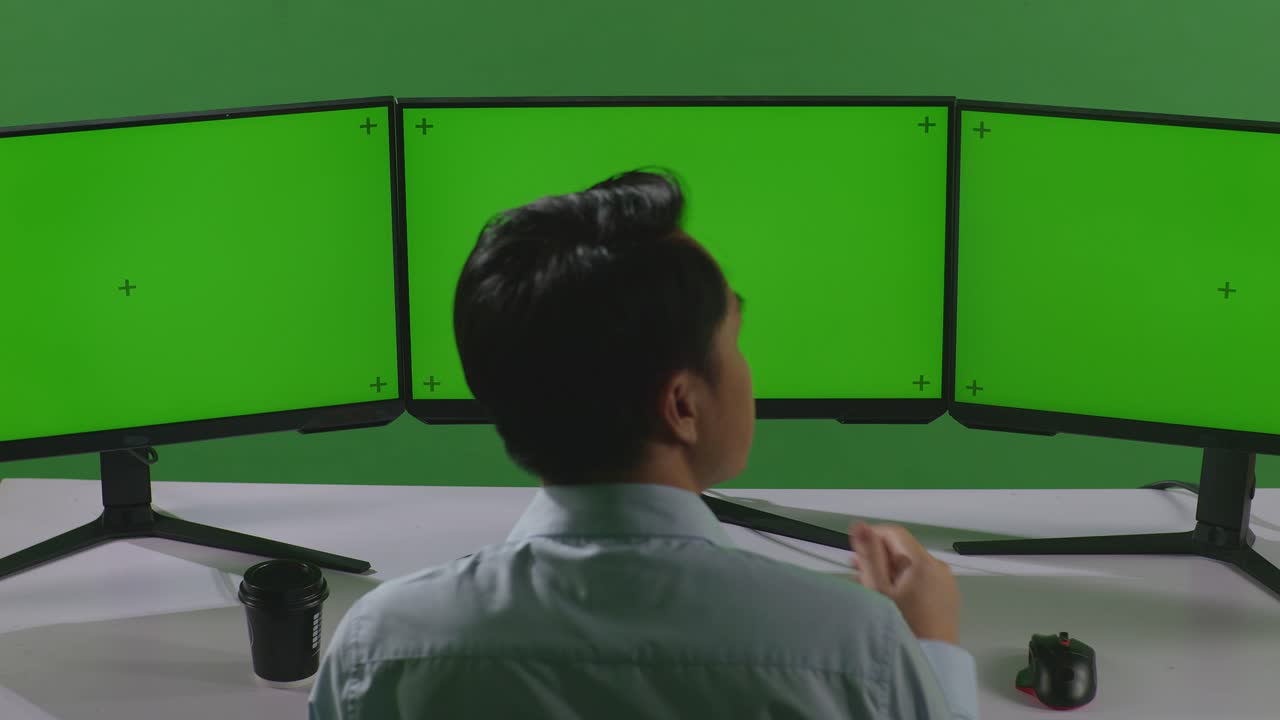 녹색 스크린 스튜디오에서 모의 여러 컴퓨터 모니터와 함께 작업하는 동안 무언가에 대해 생각하는 아시아 남자의 뒷면