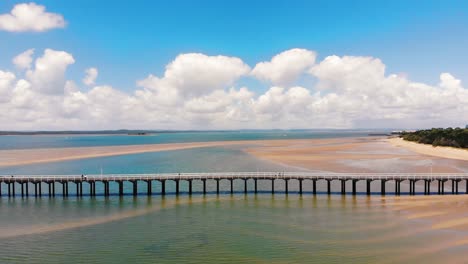 flyover of long, wooden pier stretching past camera as a wonderous, tropical world sits behind filled with gorgeous blue water, cream white sand banks and grant expanses of summer beaches on warm days