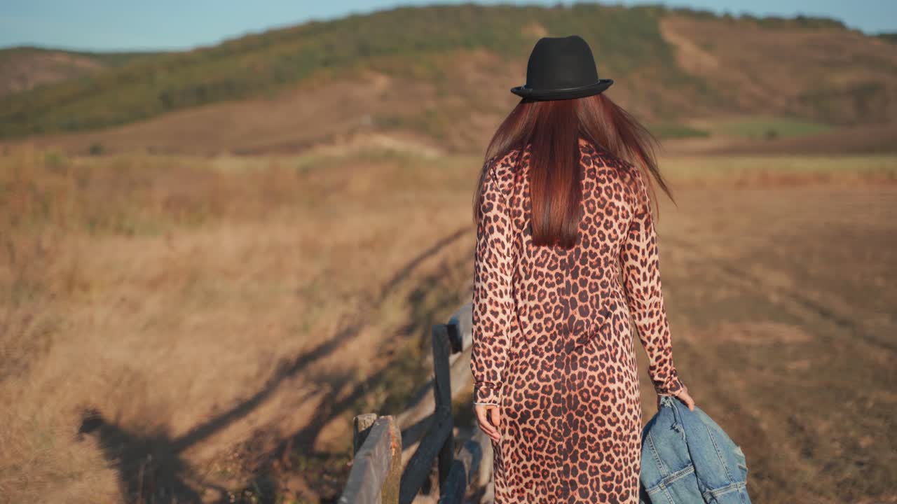 Behind view of Caucasian woman wearing fashionable printed animal dress walking and holding wooden handrail in rural flat brown countryside, handheld close up