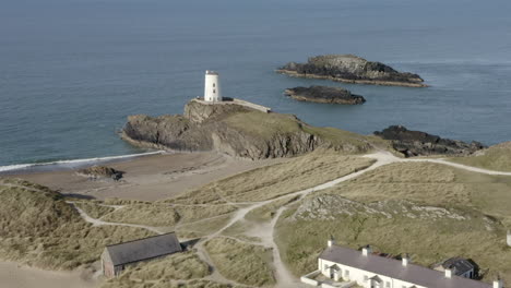 una vista aérea de la isla ynys llanddwyn que muestra las cabañas del piloto y el faro twr mawr en la distancia, volando de izquierda a derecha mientras se aleja, anglesey, norte de gales, reino unido