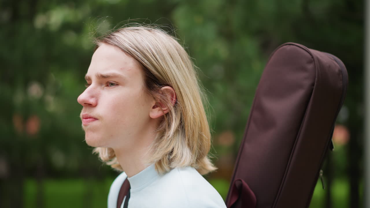 Chitarrista caucasico adolescente che cammina nel parco, si ferma nella morbida luce pomeridiana con la custodia della chitarra sulla schiena, primi piani di profilo, sfondo bokeh con foglie, espressione pensierosa, atmosfera indie lofi, casual