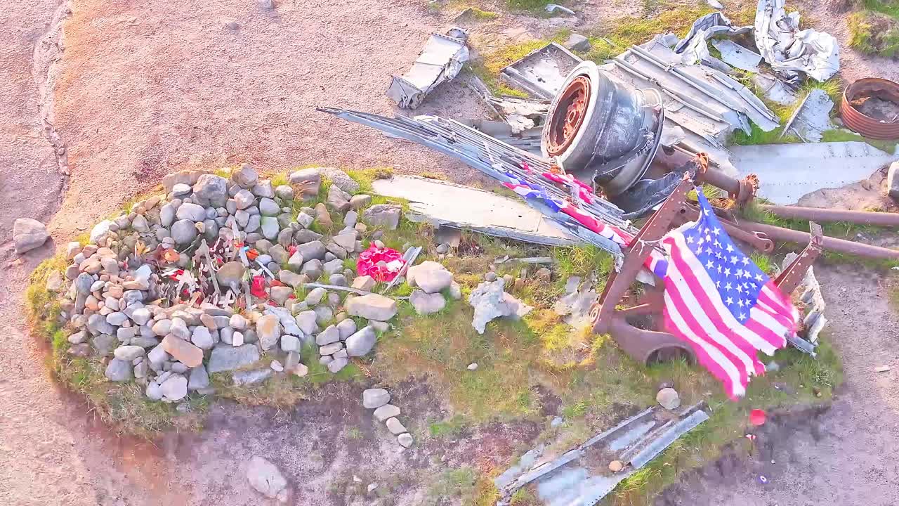 Drone rising over memorial B-29 Superfortress WWII plane wreckage site in the Peak District, England, waving US flag and stone grave in view.