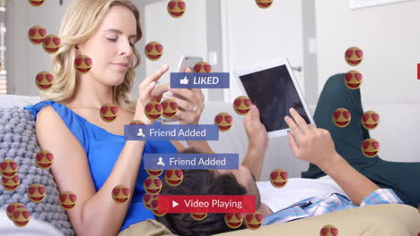 animación de ojos de corazón cara emojis flotando sobre pareja caucásica usando dispositivos electrónicos en el hogar.