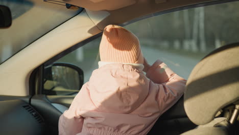 a young girl wearing a pink knit hat, white scarf, and pink jacket sits in a car, looking out the window. the scene captures her from behind as she gazes outside, highlighting a peaceful