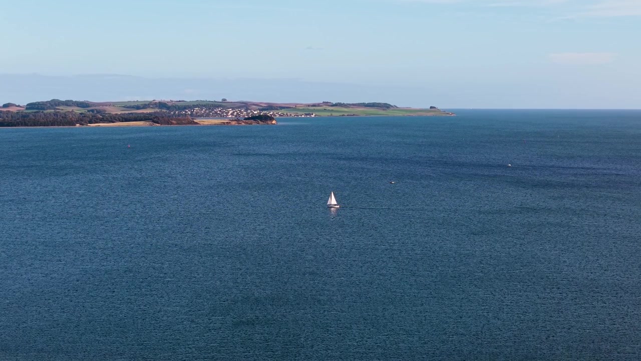Vista sull'oceano con una barca a vela in lontananza e la costa terrestre sotto un cielo sereno.