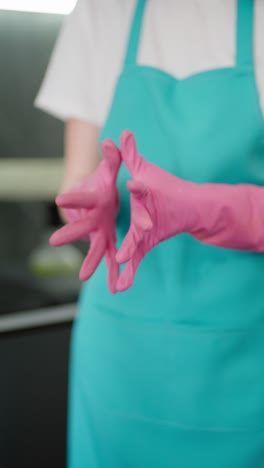 Vertikales-Video-Eines-Mädchens-In-Einer-Blauen-Schürze,-Eine-Professionelle-Reinigungskraft-Zieht-Rosa-Gummihandschuhe-An,-Bevor-Sie-Mit-Der-Reinigung-Beginnt.-In-Einer-Modernen-Wohnung-Steht-Eine-Reinigungskraft-Auf-Abruf-Bereit