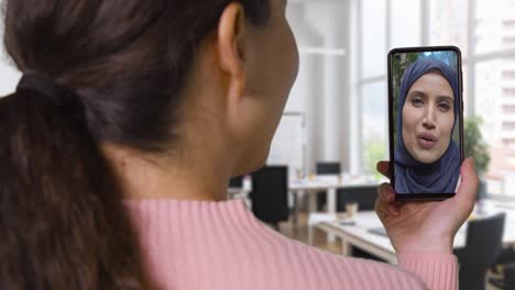 close up of businesswoman in office having video call with female muslim colleague on mobile phone