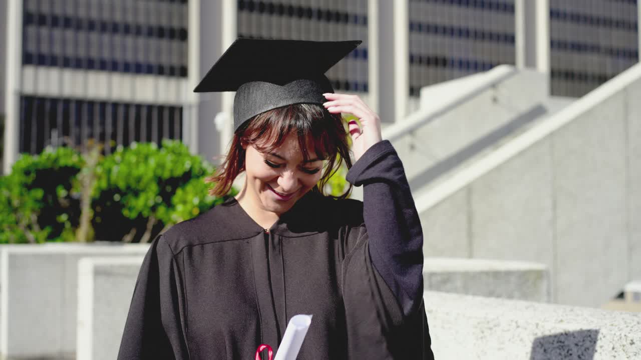 Donna di mezza età in toga e tocco neri che sorride, tenendo in mano un diploma con nastro rosso, posando per la fotocamera in una piazza.