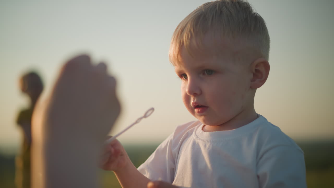 Close-up of a young boy in a white shirt, blowing bubbles from a wand while another hand is also seen blowing. A blurred figure of another child is visible in the background