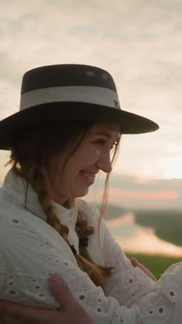 una esposa en un vestido blanco, con un sombrero y pulseras, sonríe cálidamente mientras toca amorosamente el rostro de su marido en un campo cubierto de hierba al atardecer.