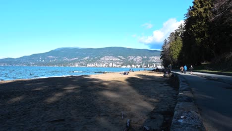 people walking and running over a road at third beach in stanley park where waves are slowly rooling and the sand on a sunny summer day