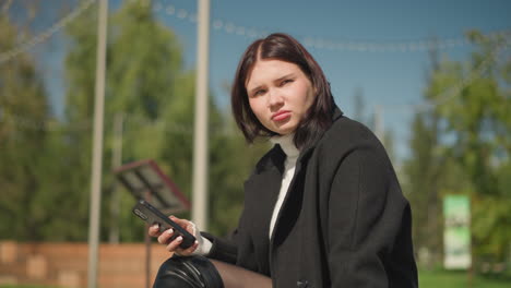 contemplative woman seated outdoors holding phone, wearing black coat and white sweater, looking thoughtfully away, with her ring-adorned hand visible, background features blurred greenery and trees