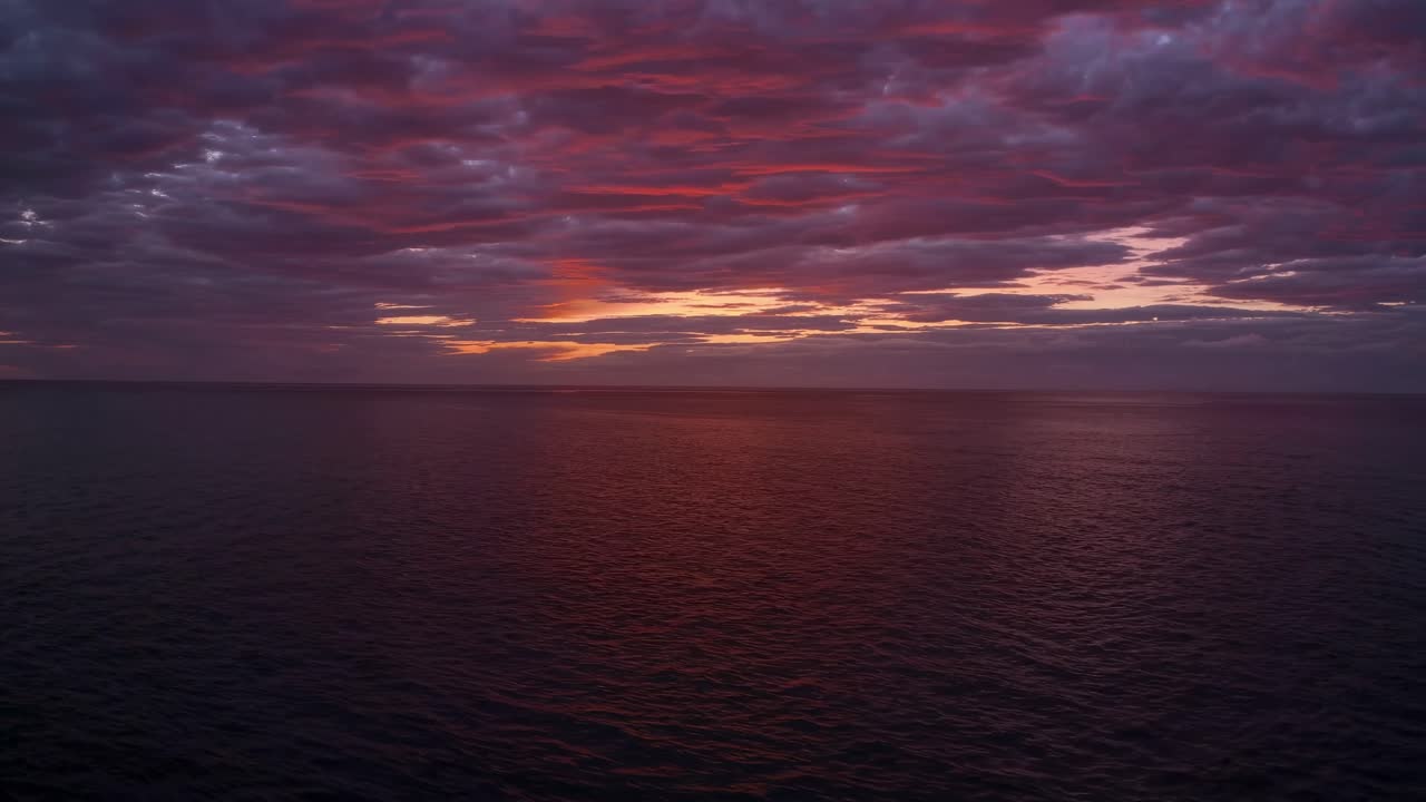 Aerial video captures a serene ocean at sunset, with vibrant pink and blue clouds
