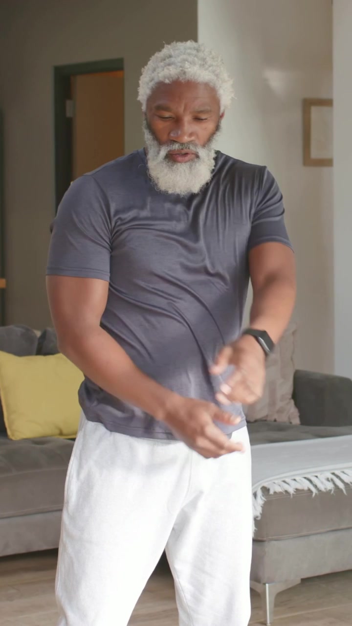 Vertical video: Pulling right arm across, man stretching and swinging at home, grey tee watch