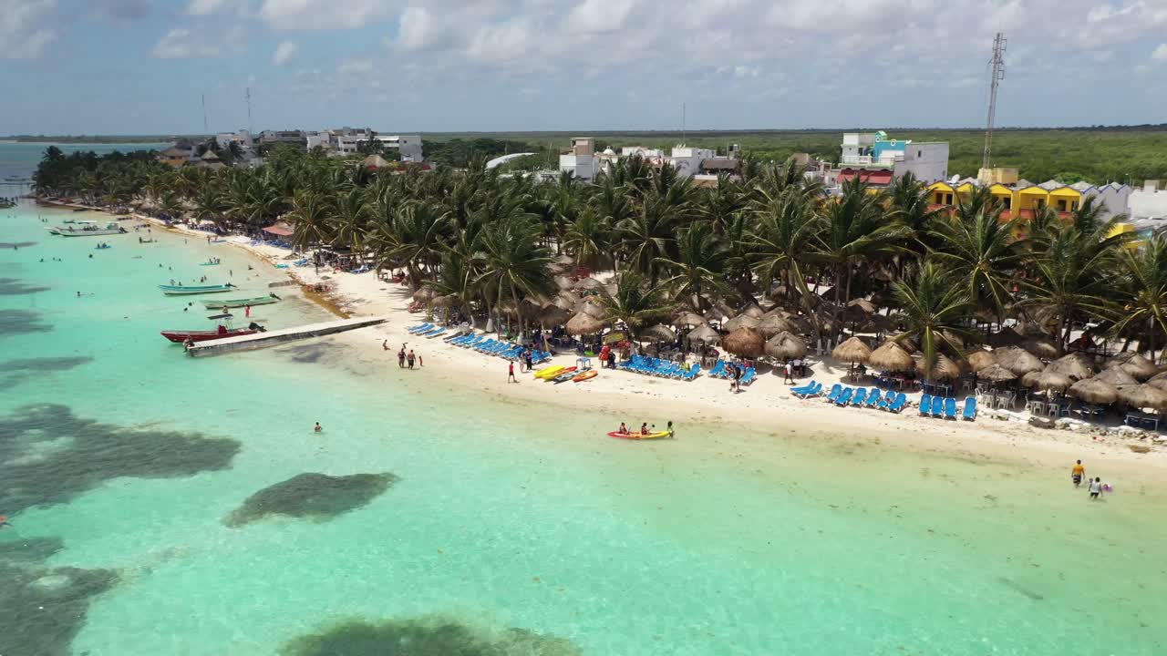 Impresionante vista aérea de Bacalar, México, con aguas cristalinas de laguna turquesa, playa bordeada de palmeras y cabañas con techo de paja. Escena de paraíso tropical con gente disfrutando de la costa caribeña