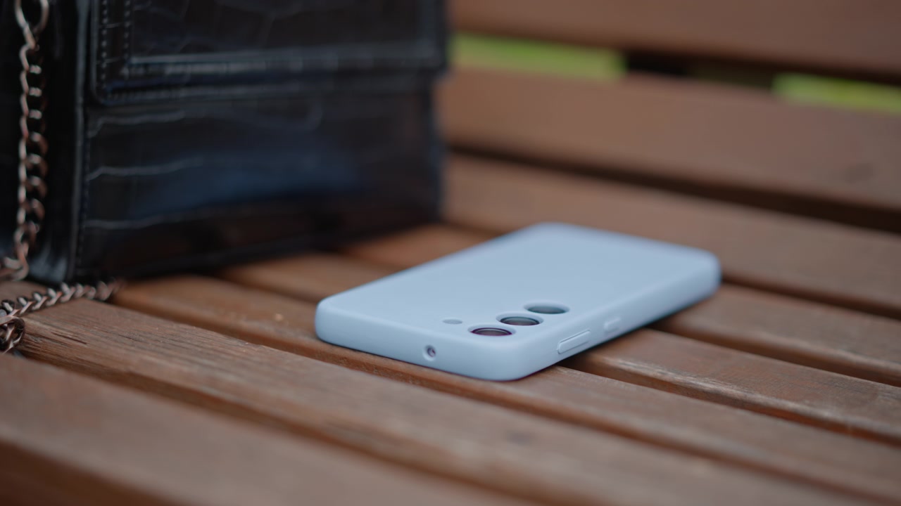 Hvid smartphone ligger med skærmen nedad med en let hældning på en træbænk med lameller ved siden af en sort lædertaske med metalkæderem; et lille mellemrum antyder et nyligt skred og en stille udendørs urban pause