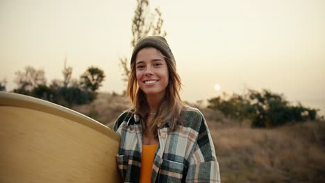 Portrait-of-a-happy-blonde-girl-in-a-hat-in-a-plaid-shirt-who-is-holding-a-wooden-surfboard-in-her-hands-smiling-and-looking-at-the-camera-against-the-backdrop-of-the-sea-and-trees-and-bushes-on-the-slope-of-the-shore