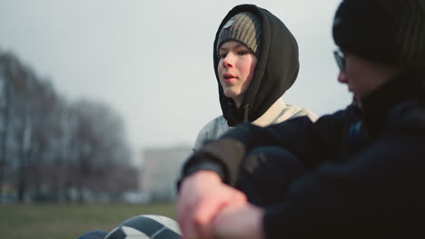 close-up of two boys seated on a grassy field, with one in gray clothing holding and playing with a soccer ball