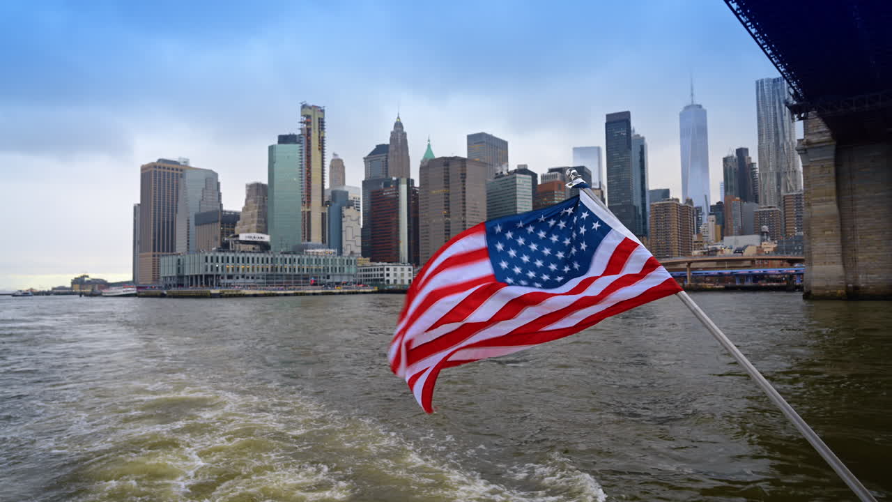 Bandiera statunitense su barca con vista sullo skyline di Lower Manhattan. Bandiera a stelle e strisce che sventola su una barca con il One World Trade Center e il Ponte di Brooklyn in lontananza.