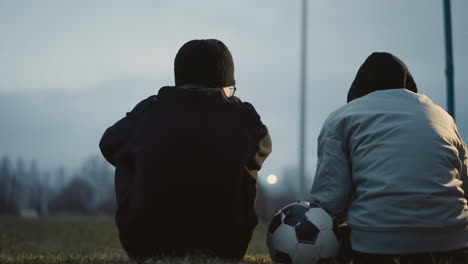 two boys are seated closely together on a grassy field at dusk, with one resting his hand on a soccer ball, with a blur view of someone passing by in the background