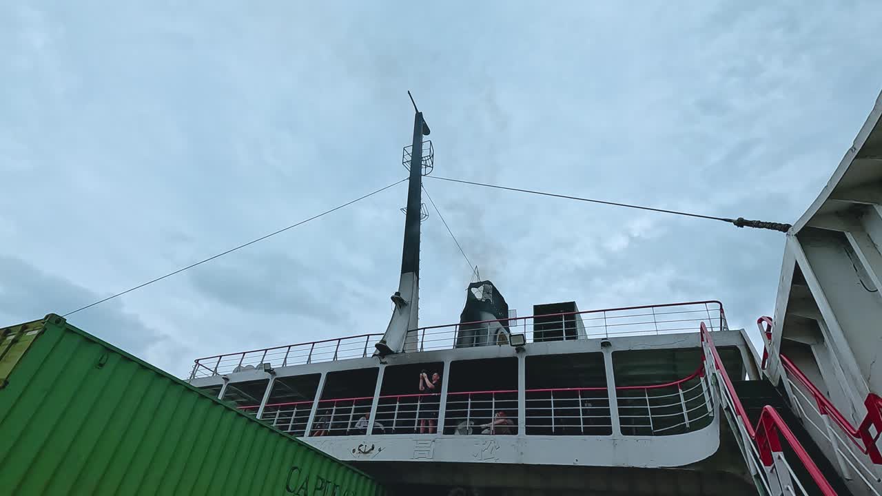 A truck with a green container boards a ferry under cloudy skies in Koh Samui, Thailand. The scene captures movement and industrial activity
