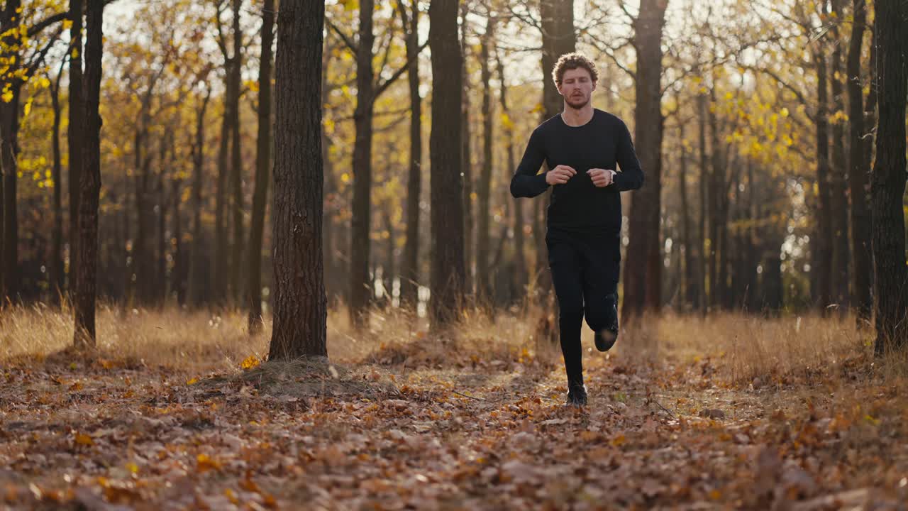 um homem com cabelos encaracolados e uma barba em um uniforme esportivo moreno preto olha para o relógio e, em seguida, corre rapidamente para o seu destino durante o seu sprint e jogging de manhã na floresta de outono
