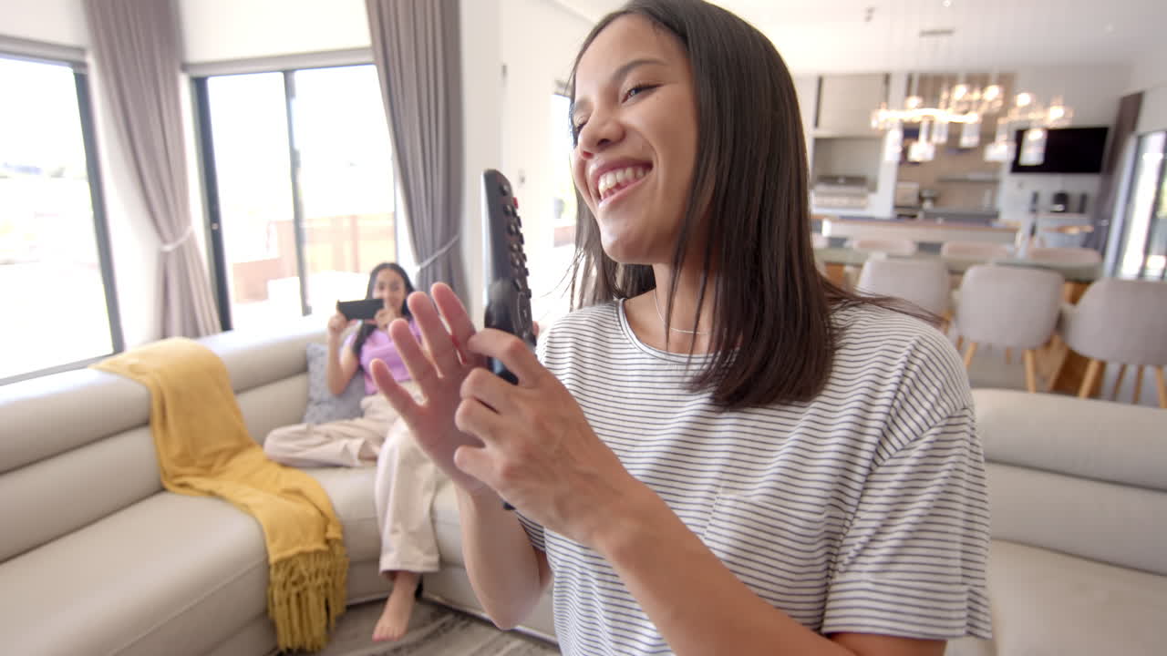Teenager holding TV remote while friend relaxing on couch using smartphone